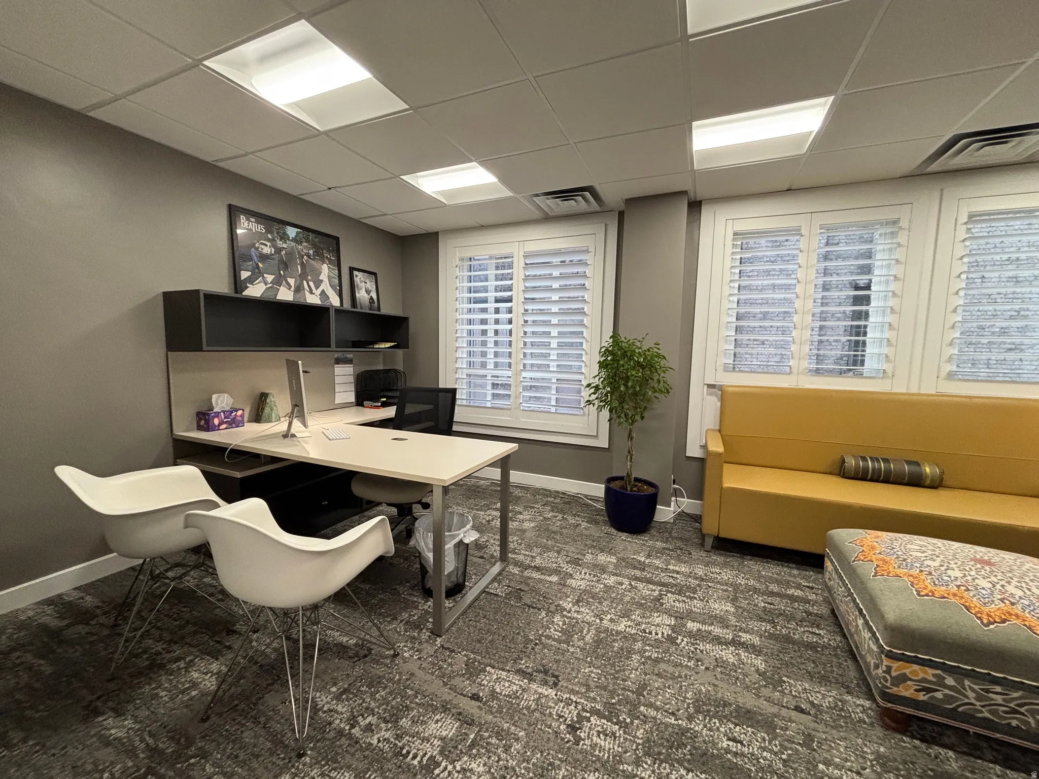 Professional office space featuring three windows with white plantation shutters, suspended ceiling with recessed lighting, integrated desk with shelving, and patterned carpet flooring