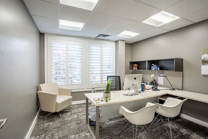 Office space featuring plantation shutters on multiple windows, textured carpeting, a suspended acoustic tile ceiling with integrated lighting, and built-in shelving