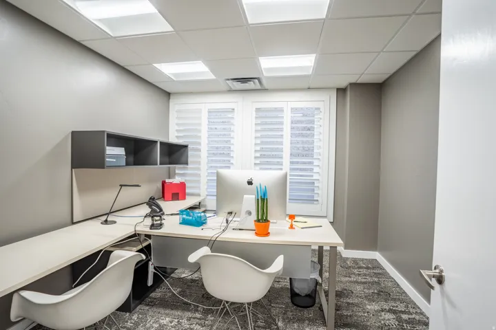 Dedicated office space featuring a large L-shaped desk with integrated shelving, shuttered windows, recessed ceiling lighting, and patterned carpet flooring