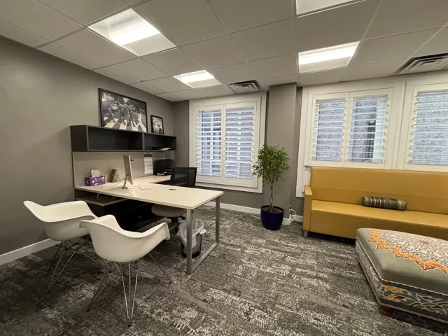 Professional office space featuring three windows with white plantation shutters, suspended ceiling with recessed lighting, integrated desk with shelving, and patterned carpet flooring