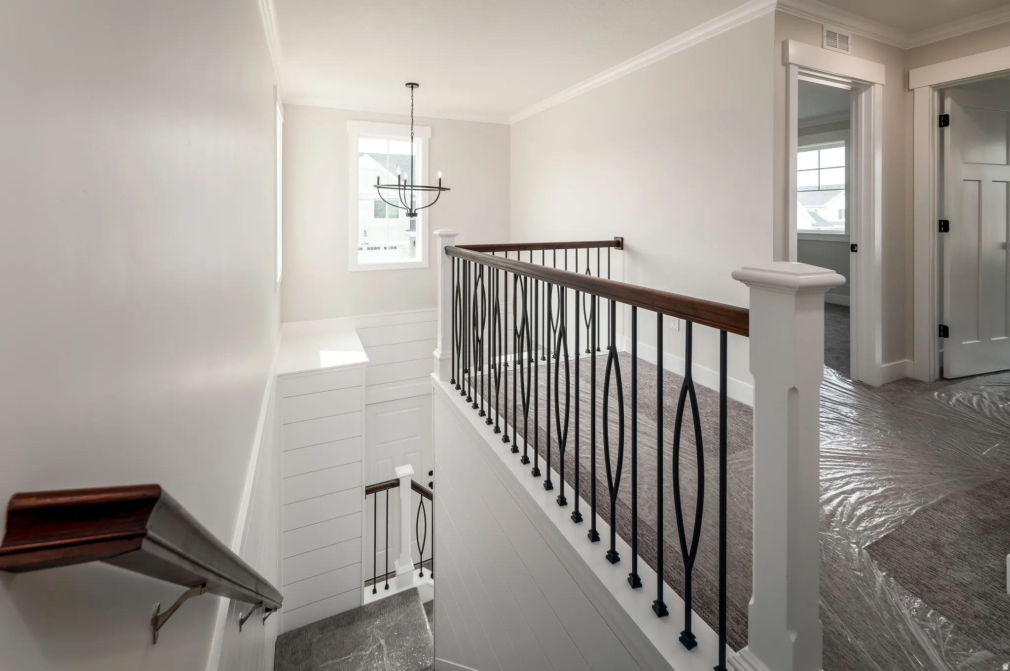Upper landing featuring a decorative wrought iron and wood-finish railing, a prominent window, a contemporary chandelier, crown molding, and a paneled wall detail along the staircase
