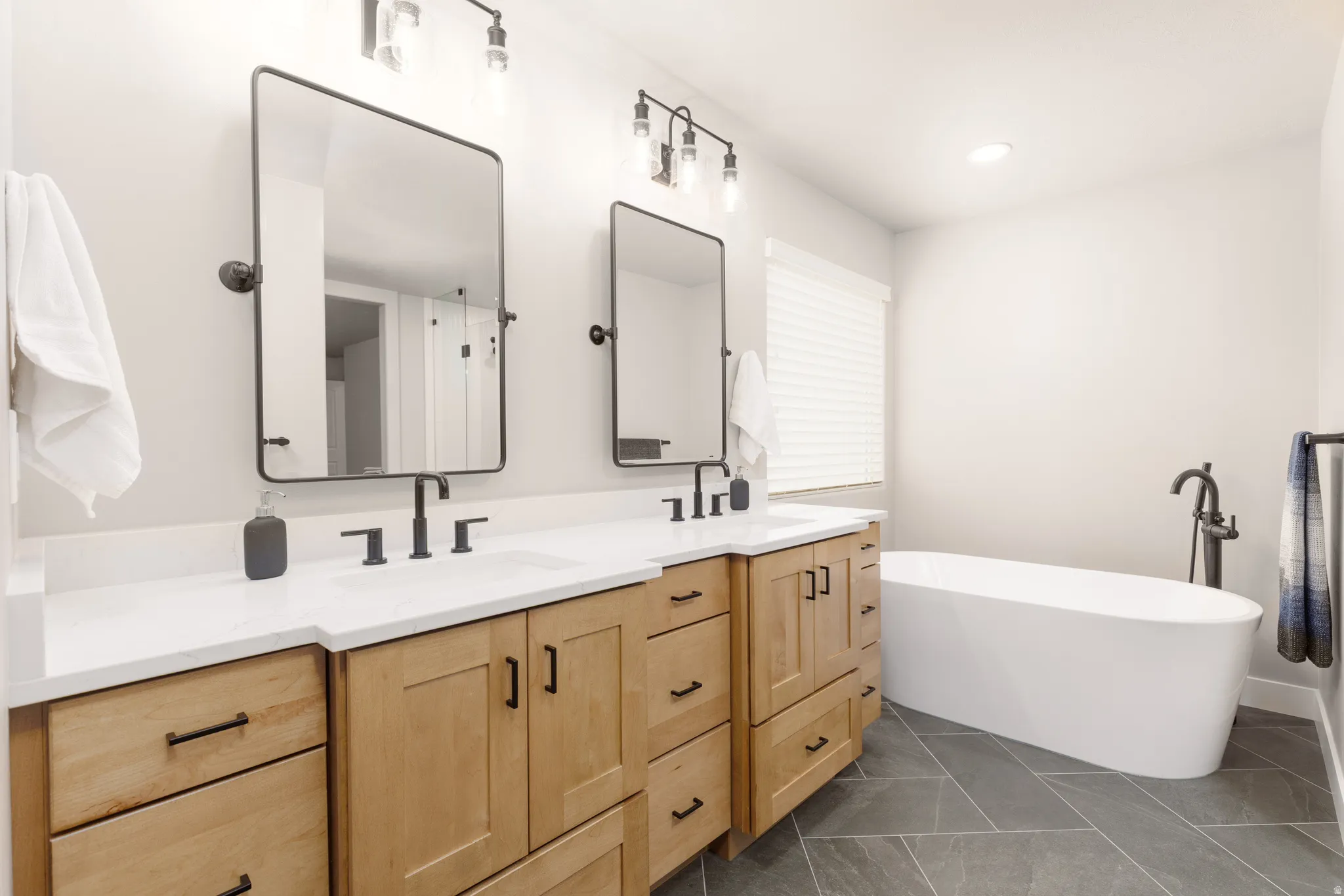Primary ensuite featuring a freestanding soaking tub with a floor-mounted faucet, dual vanity with a white countertop and wood-finish cabinetry, a rectangular mirror with a black frame, and a shower with patterned accent tiling