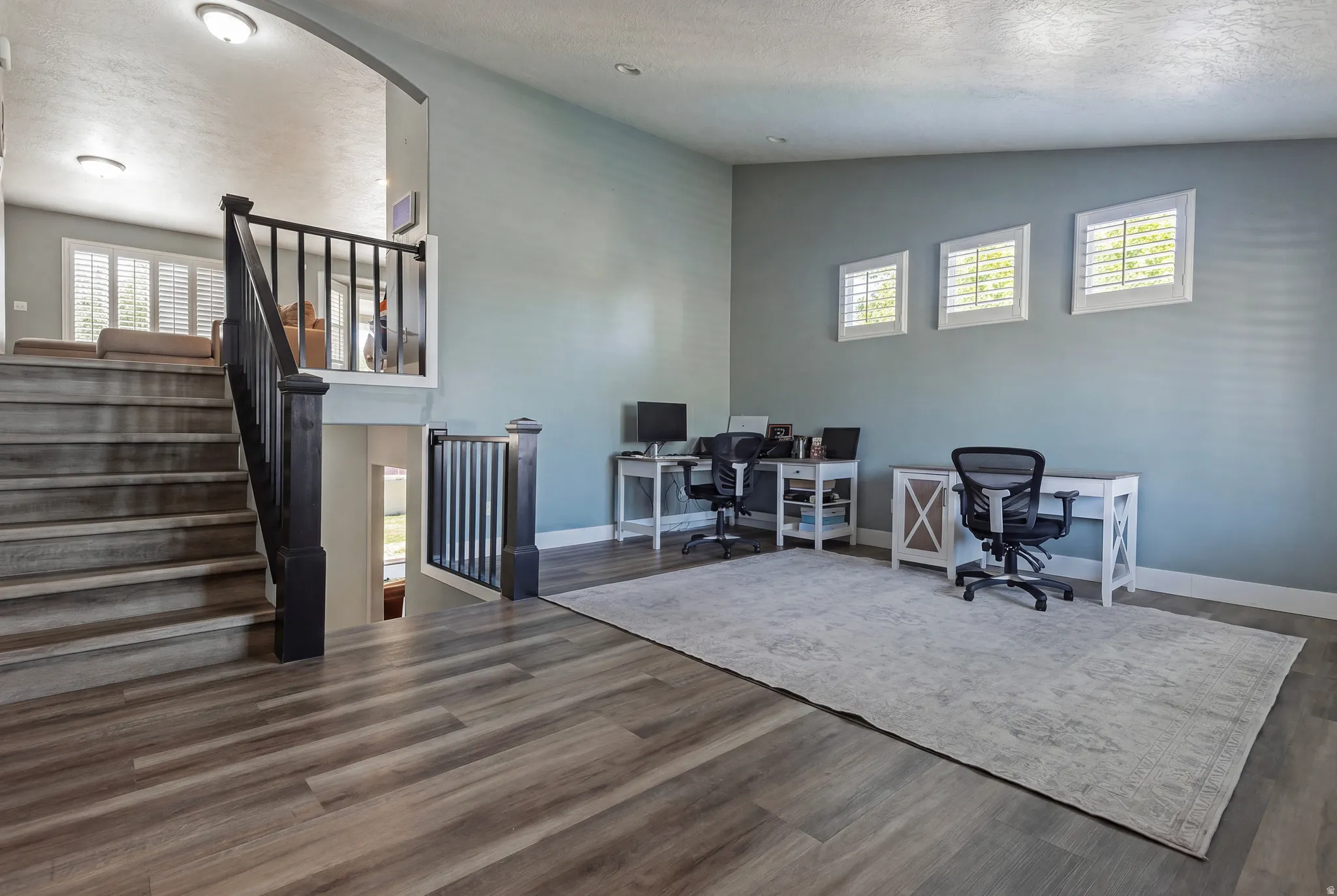Spacious open-concept area featuring wood-finish flooring, a contemporary staircase with dark wood newel posts and spindles, three windows with plantation shutters, and light blue wall paint