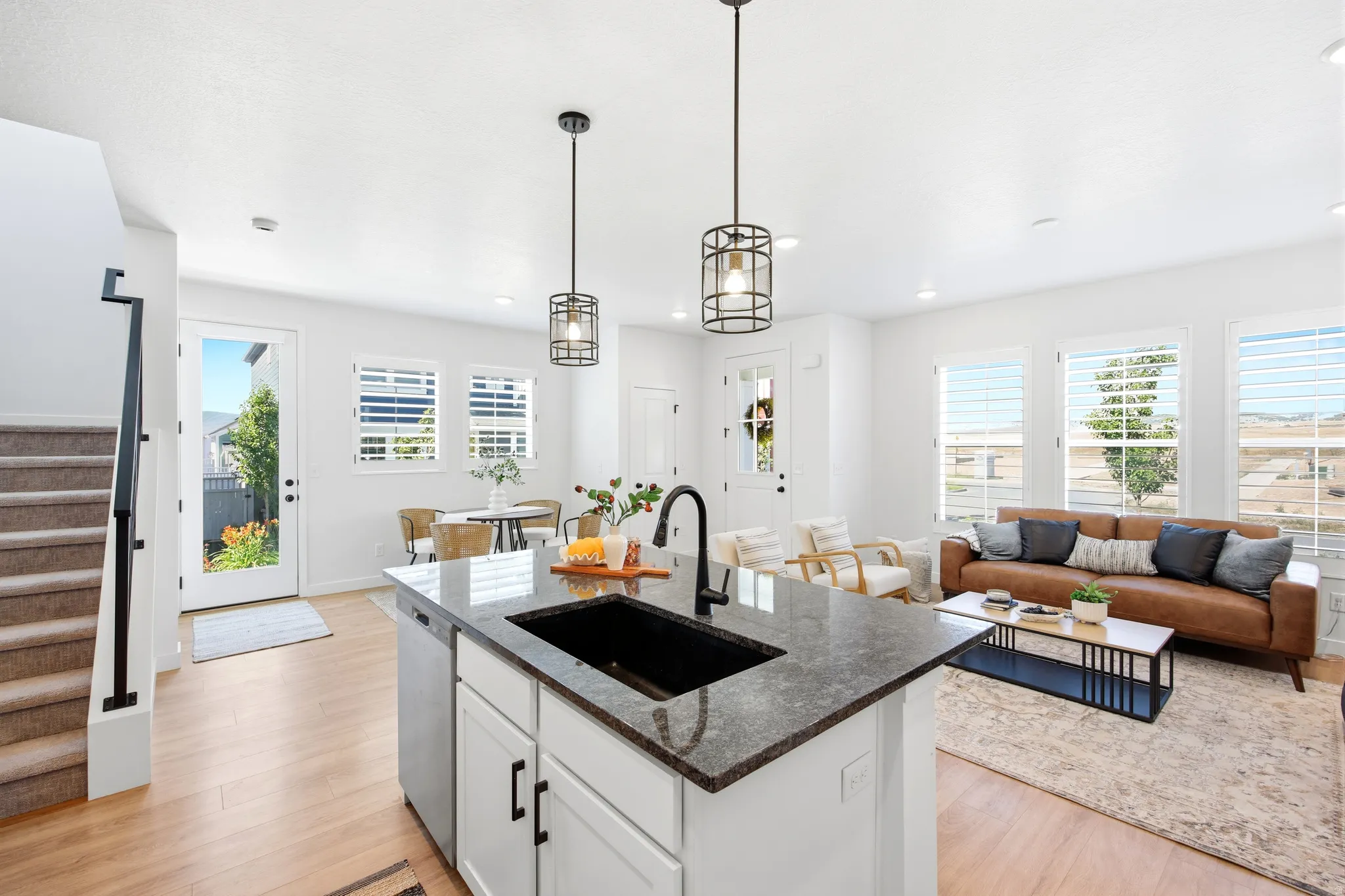 Open-concept living space featuring a central kitchen island with a dark countertop and undermount sink, pendant lighting, light wood-finish flooring, and multiple windows with plantation shutters