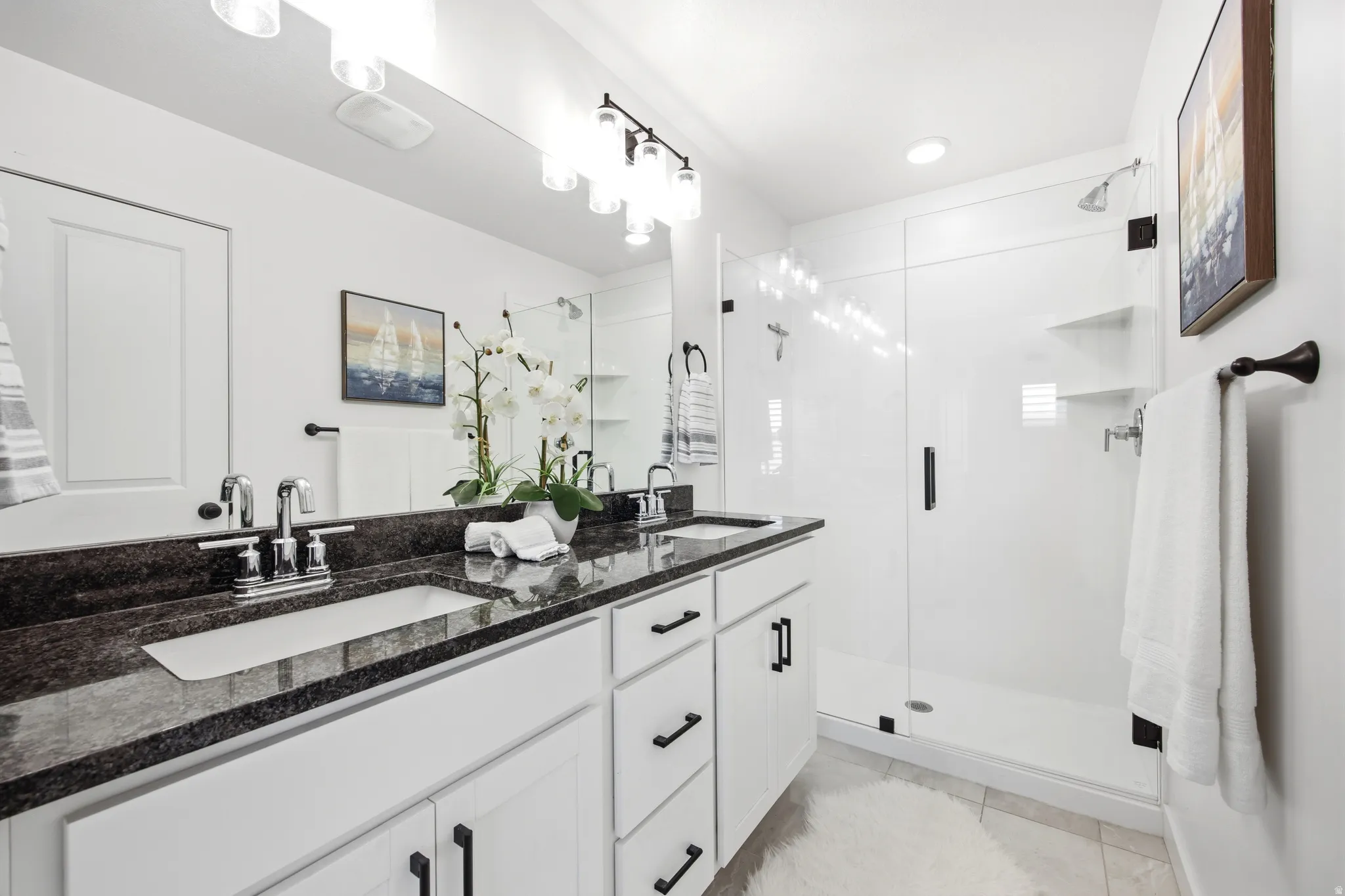 Modern bathroom featuring a dual vanity with a dark granite countertop, white cabinetry with black hardware, and an expansive mirror