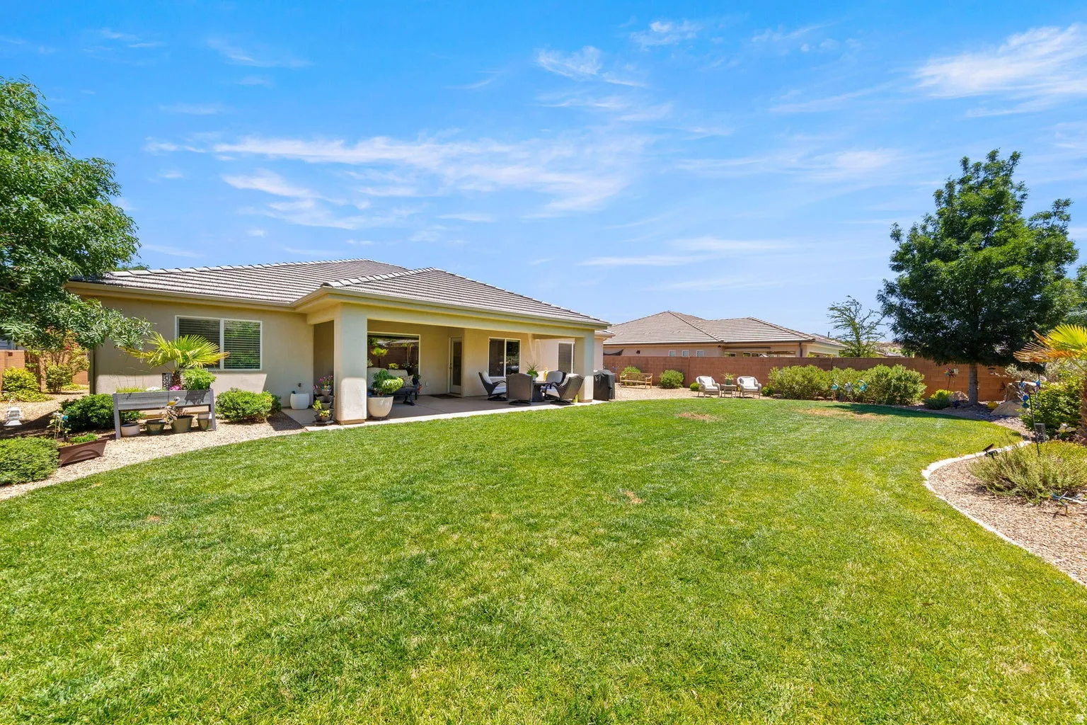 Expansive green lawn with a curved landscape bed, mature trees, a covered patio, and stucco exterior