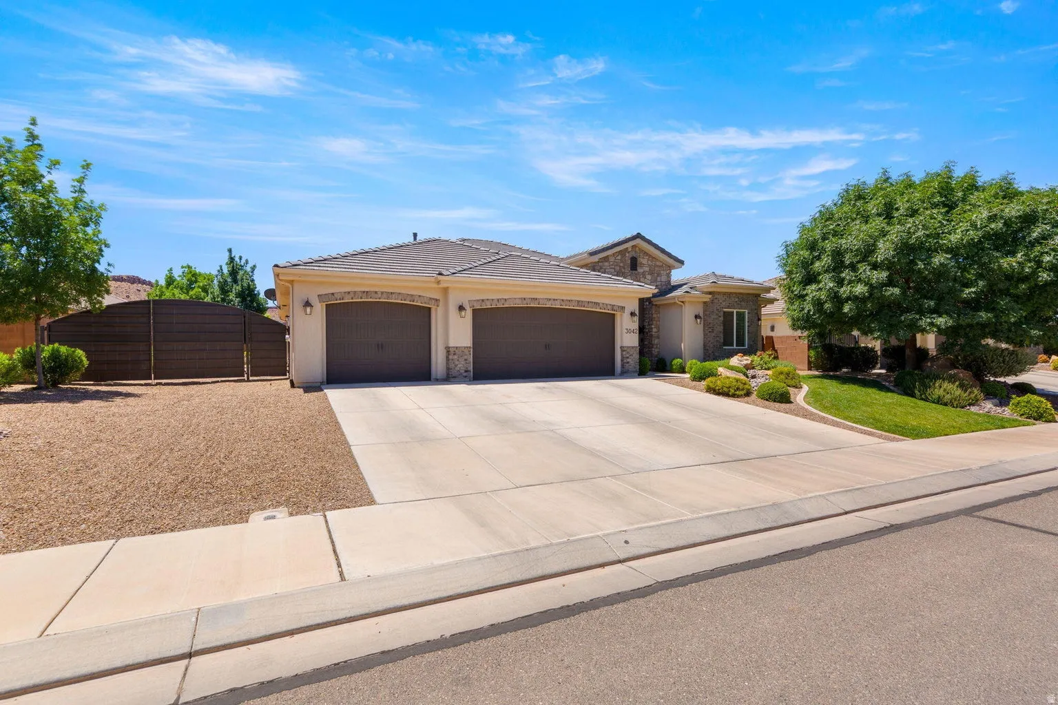 Contemporary stucco and stone-veneer exterior featuring a two-car garage with arched accents, a concrete driveway, and low-maintenance landscaping with mature trees and shrubs