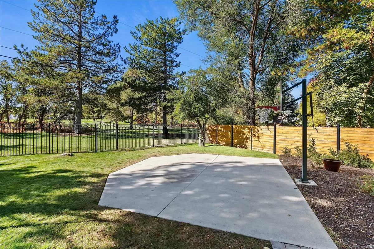 Expansive outdoor space featuring a paved concrete pad, a black metal fence, mature trees, and a horizontal plank wood fence