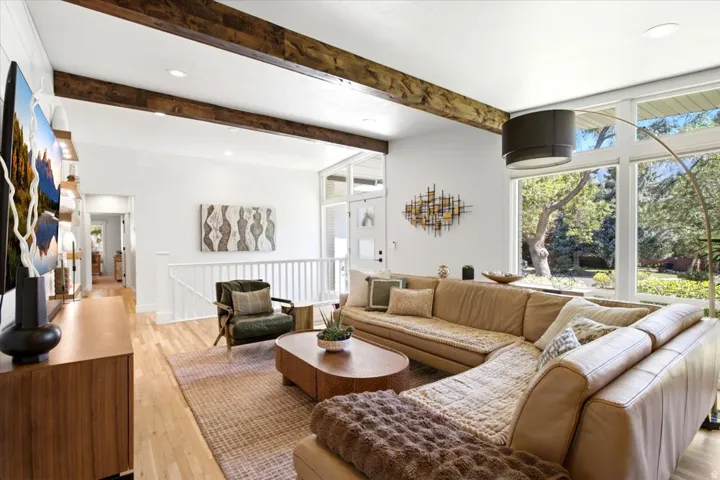 Bright living area featuring exposed wood beams, light wood-finish flooring, and expansive windows