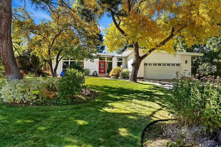 Single-story residence featuring a white vertical siding exterior, a red entry door, a two-car garage, and a lush front lawn with mature trees