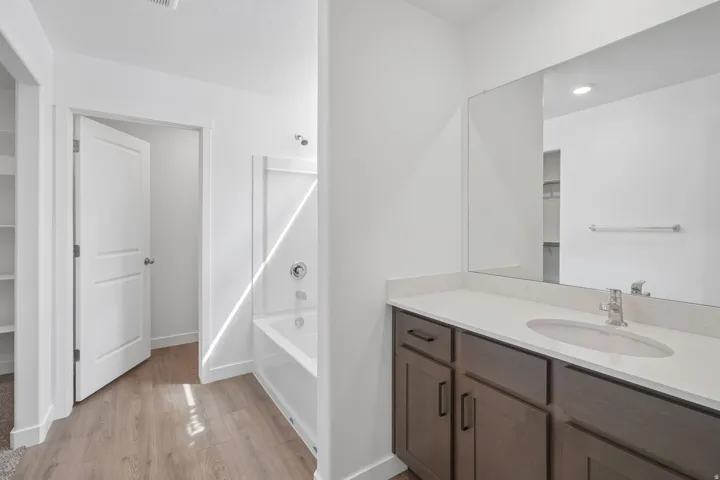 Wood-finish flooring, white vanity with integrated sink, sleek chrome faucet, large vanity mirror, and a built-in tub with shower