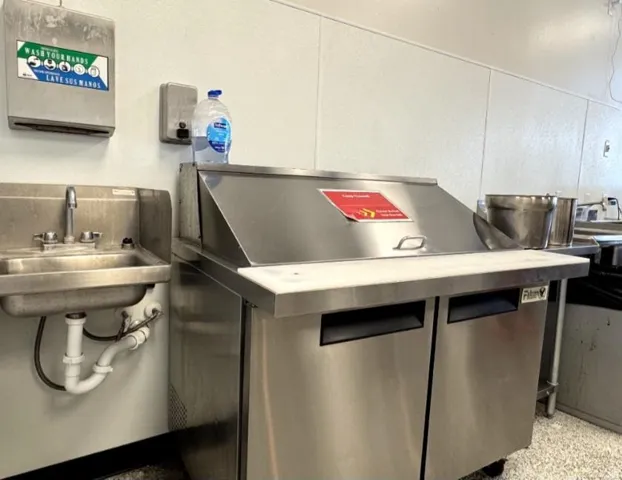 Stainless steel commercial kitchen preparation area featuring an integrated sink, wall-mounted paper towel dispenser, and a refrigerated prep station with a cutting board