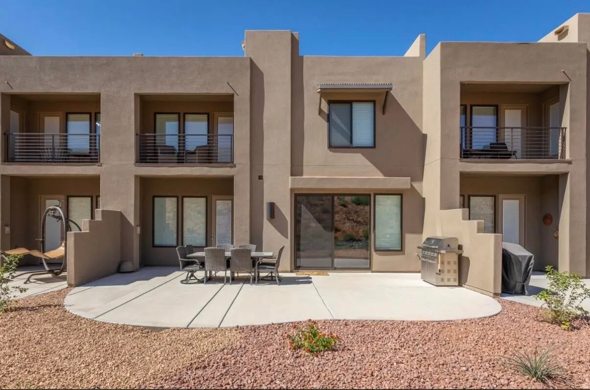 Stucco exterior featuring multiple private balconies with metal railings, a ground-level patio, and desert landscaping with rock ground cover