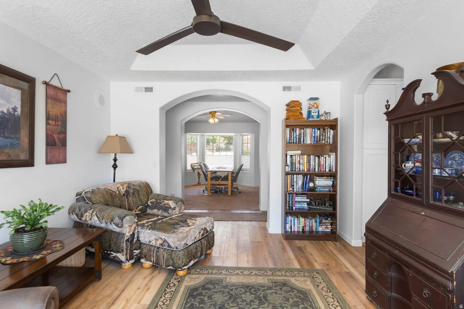 Living area featuring wood-finish flooring, a tray ceiling with a dark-bladed ceiling fan, and arched doorways
