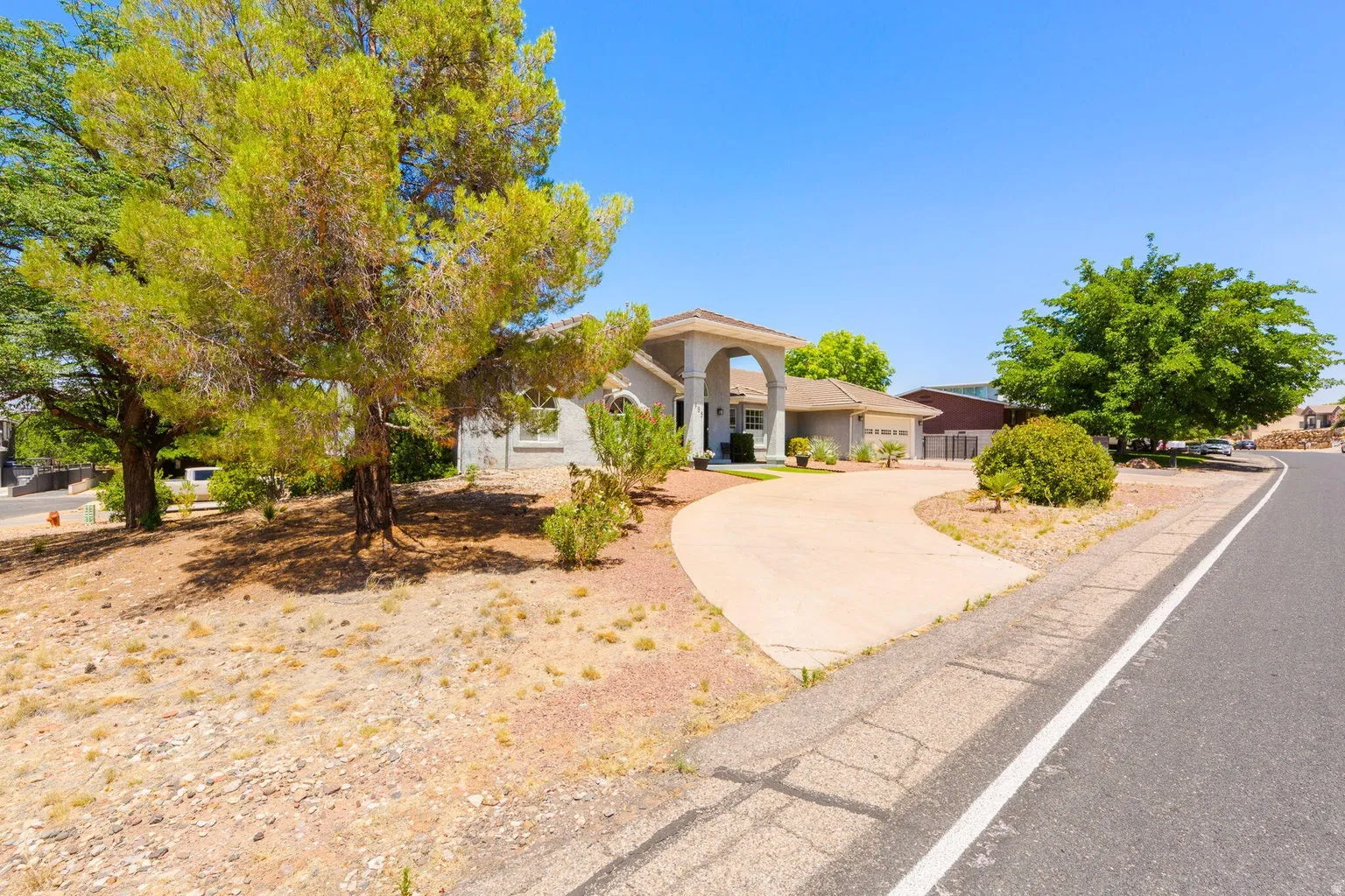 Traditional stucco exterior with a tiled roof, featuring an arched portico entryway, an attached two-car garage, mature trees, and a concrete driveway