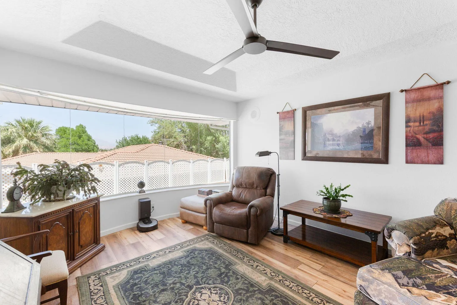 Wide window providing exterior views, wood-finish flooring, ceiling fan, white wall finishes, and a decorative console cabinet
