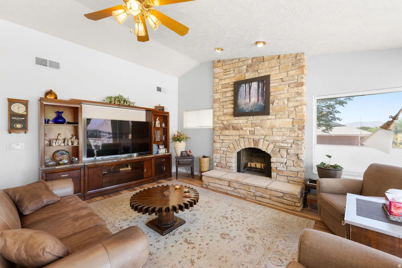 Living area featuring a floor-to-ceiling stone fireplace with an arched firebox and hearth, vaulted ceilings, a built-in media center with cabinetry, wood-finish flooring, and a large window with horizontal blinds