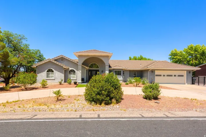 Single-story stucco residence featuring an arched portico entry, a two-car garage, arched windows, and a tiled roof, surrounded by desert landscaping and mature trees
