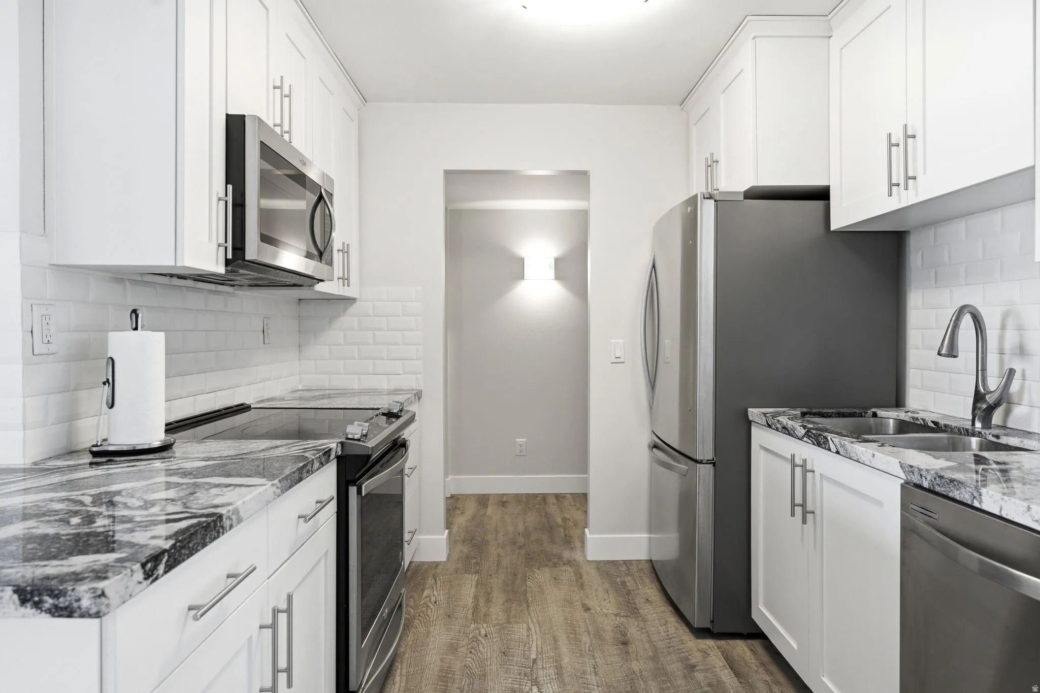 Galley kitchen featuring white shaker cabinetry, stainless steel appliances, white subway tile backsplash, wood-finish flooring, and stone countertops