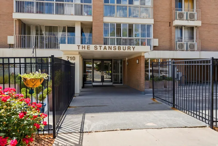 Building entrance featuring a brick facade, glass double doors, a prominent "THE STANSBURY" sign, and metal fencing