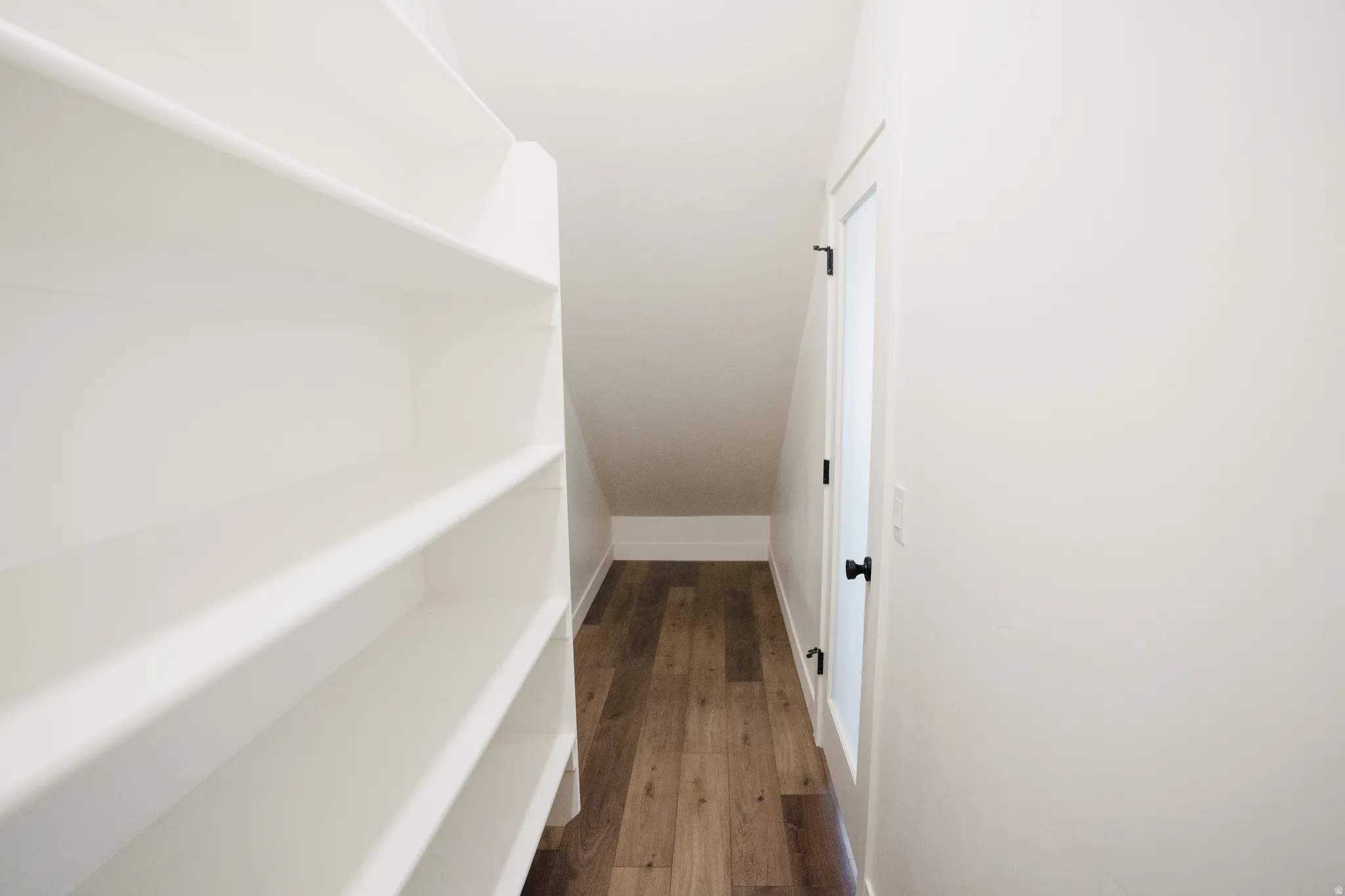 Angled storage room featuring extensive built-in shelving, wood-finish flooring, white walls, and a white door with matte black hardware