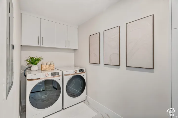 Dedicated laundry area featuring overhead white cabinetry with silver hardware, light-toned wall paint, and white flooring with subtle veining