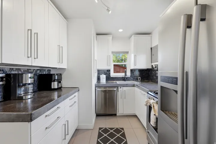Contemporary kitchen featuring white cabinetry, stainless steel appliances, dark concrete  countertops, and a decorative mosaic tile backsplash, and light tile flooring