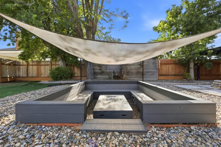 Exterior fire pit area featuring built-in composite benches, a shade sail, and surrounding river rock landscaping