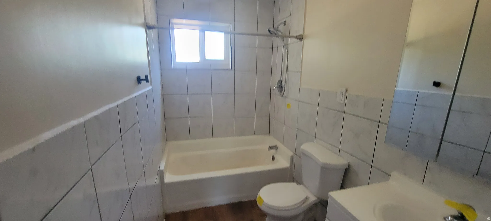 Bathroom featuring white wall tile, a white bathtub with shower fixture, a window with frosted glass, and wood-finish flooring
