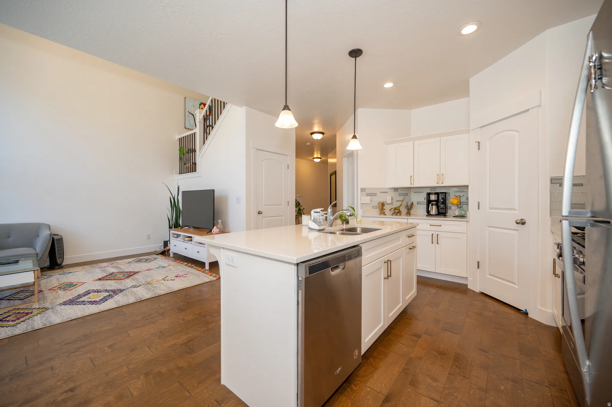 Open-concept living area featuring wood-finish flooring, a kitchen island with integrated sink, white cabinetry, stainless steel appliances, and a tiled backsplash