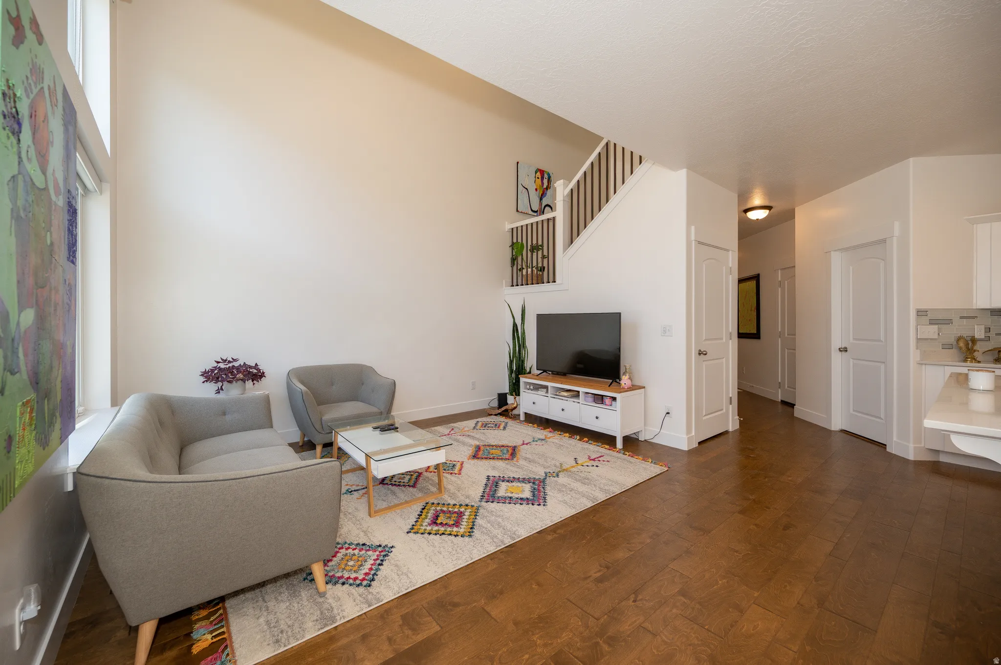 Living room featuring a soaring vaulted ceiling, rich wood-finish flooring, a white staircase with wood railings, and a partial view of a kitchen with white cabinetry and a tiled backsplash
