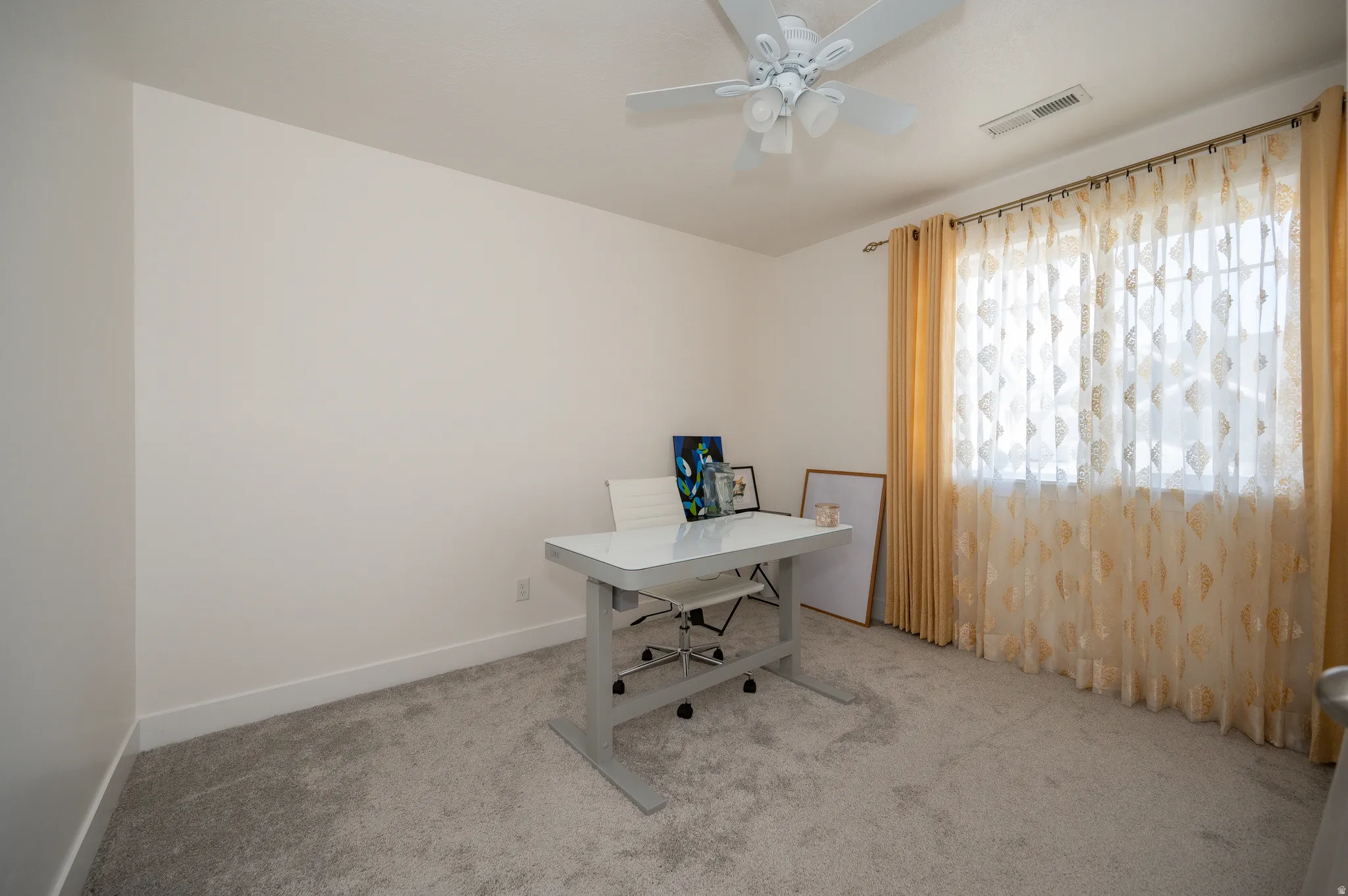 Room featuring light neutral wall paint, gray carpeting, a window with curtain rod and sheer and opaque drapes, white baseboards, and a white ceiling fan with light fixture