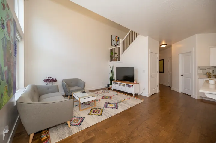 Living room featuring a soaring vaulted ceiling, rich wood-finish flooring, a white staircase with wood railings, and a partial view of a kitchen with white cabinetry and a tiled backsplash