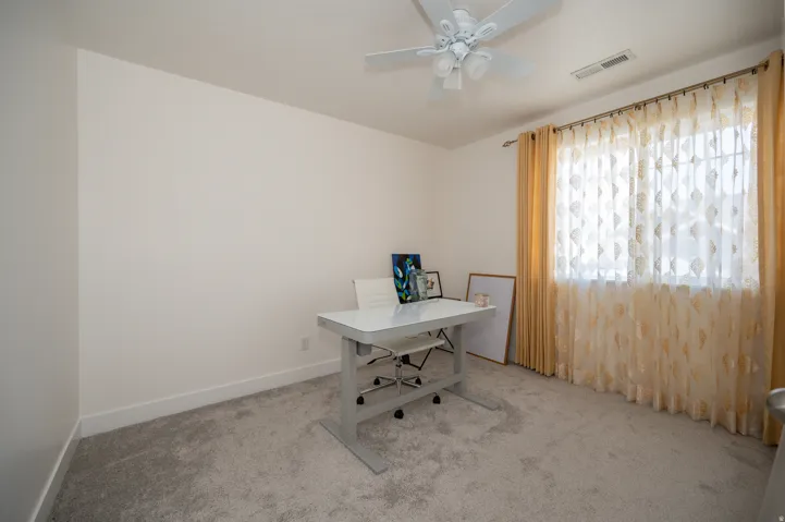 Room featuring light neutral wall paint, gray carpeting, a window with curtain rod and sheer and opaque drapes, white baseboards, and a white ceiling fan with light fixture