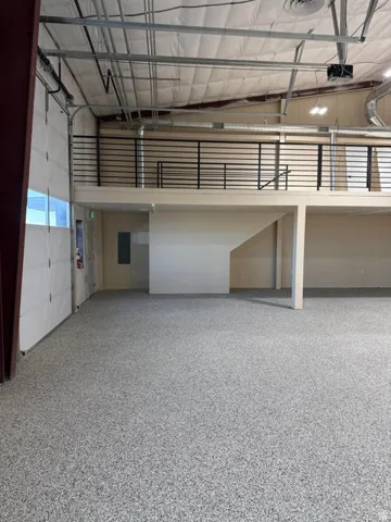 Spacious interior featuring a large overhead door, an exposed metal ceiling with ductwork, and a second-level mezzanine with a black metal railing