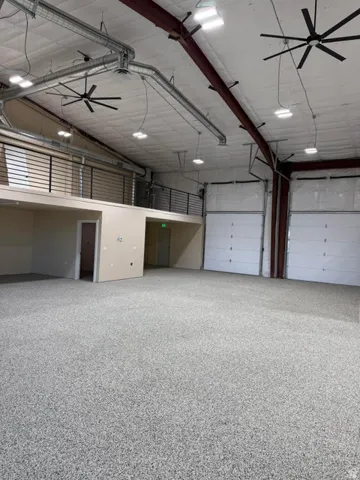 Spacious interior featuring a large open floorplan with a textured epoxy floor, two overhead garage doors, an upper-level mezzanine with a black metal railing, exposed ductwork, and multiple ceiling fans