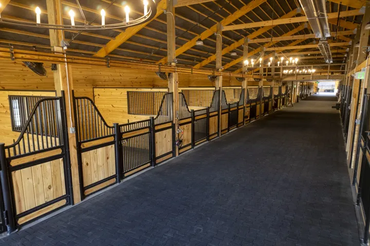 Barn interior featuring multiple stalls with wood and black metal construction, exposed wood beam ceiling, overhead lighting, and a dark-toned textured floor