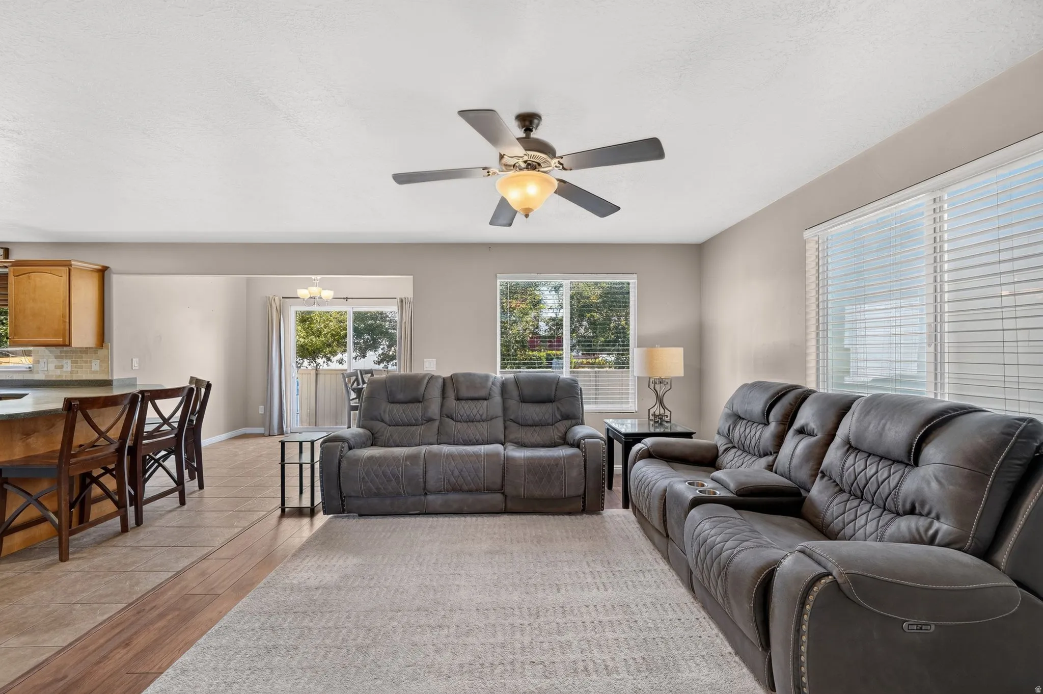 Expansive main living area featuring a ceiling fan with integrated lighting, multiple windows with blinds, a sliding glass door, and a combination of wood-finish flooring and tile