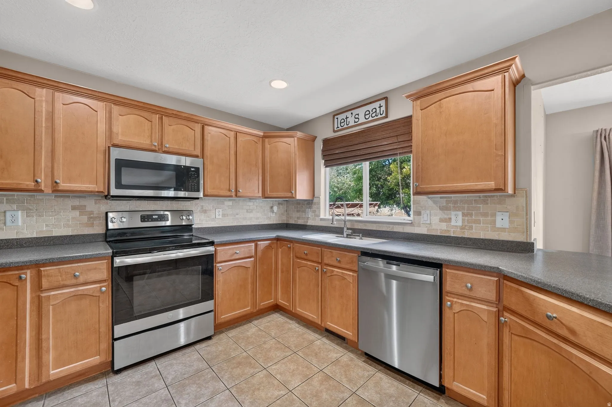Kitchen featuring wood cabinetry, stainless steel appliances, a tile backsplash, dark countertops, and tile flooring