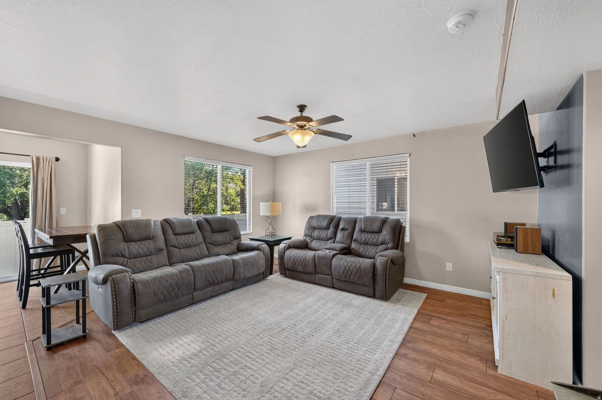 Open-concept living area featuring wood-finish flooring, a ceiling fan with integrated lighting, and multiple windows