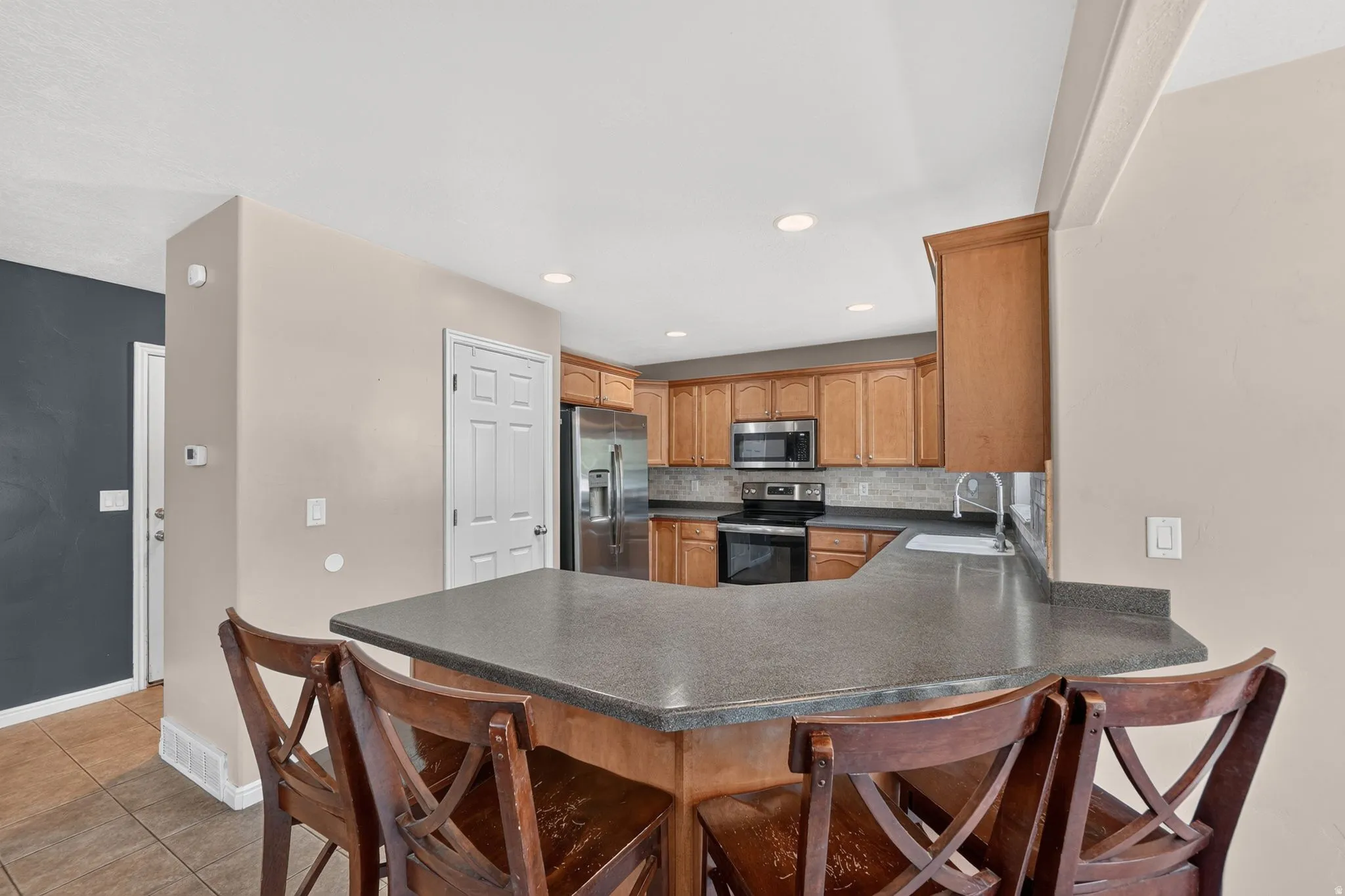 Kitchen featuring a U-shaped breakfast bar with a stone-finish countertop, recessed lighting, wood-finish cabinetry, stainless steel appliances, and a tile backsplash