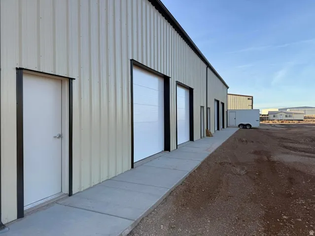 Industrial-style exterior with corrugated metal siding, multiple white roll-up garage doors, a pedestrian door, a concrete walkway, and an expansive gravel lot