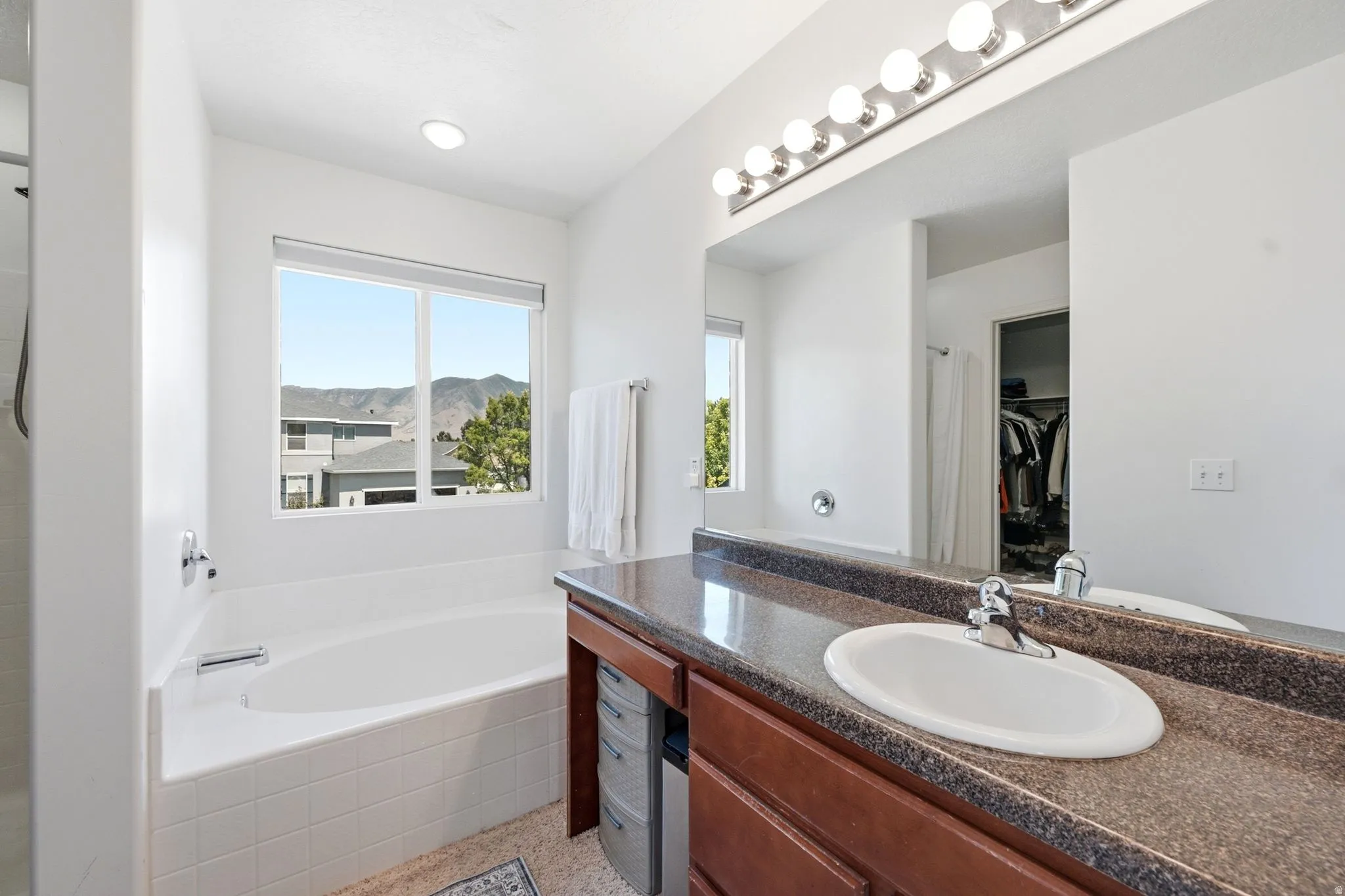 Spacious bathroom featuring an oval soaking tub with tile surround, a large window overlooking a mountain landscape, a wood-finish vanity with an oval undermount sink, a stone-finish countertop, and a wide mirror with overhead lighting