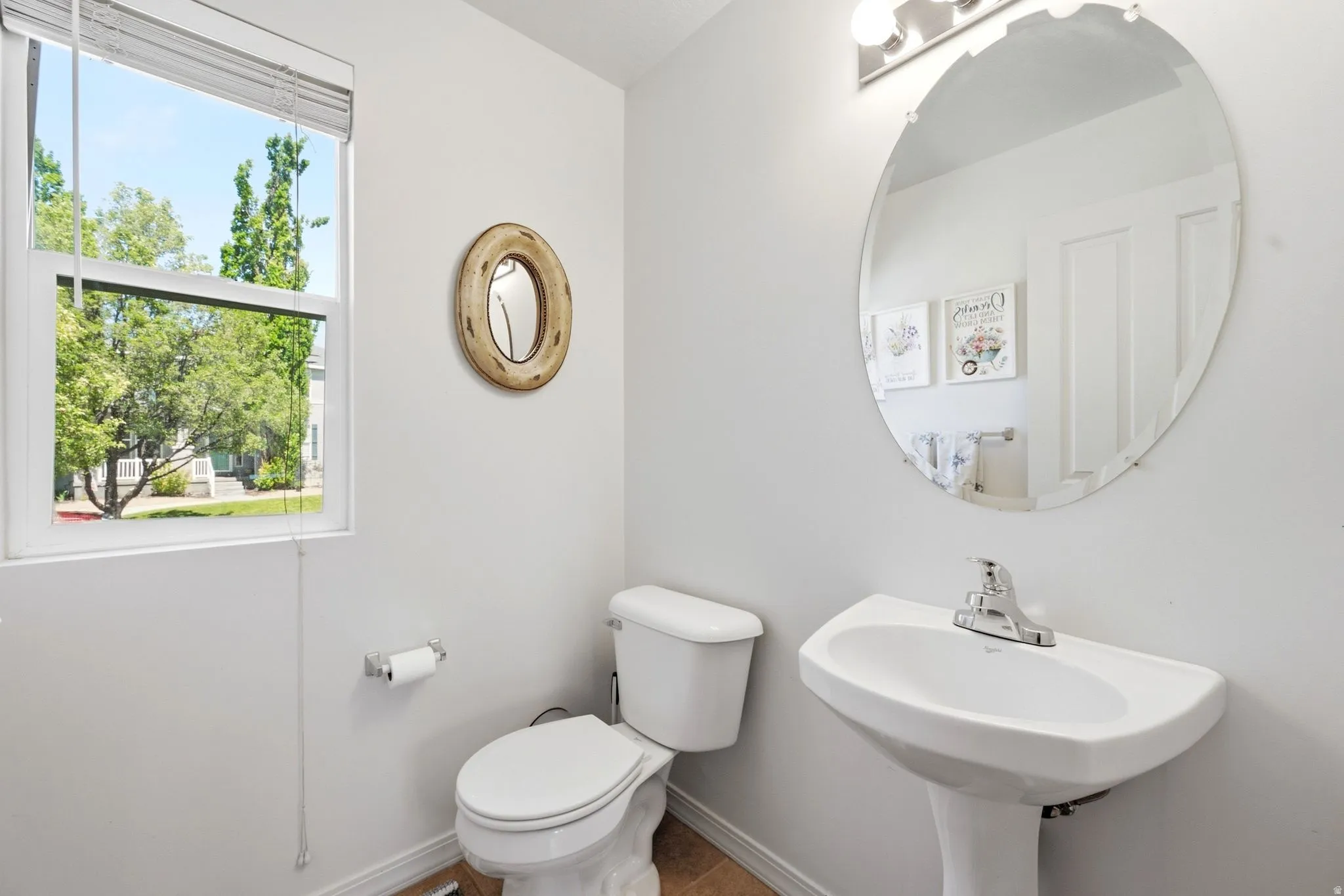 Pedestal sink featuring a chrome faucet, oval mirror, window with privacy blinds, and light tile flooring