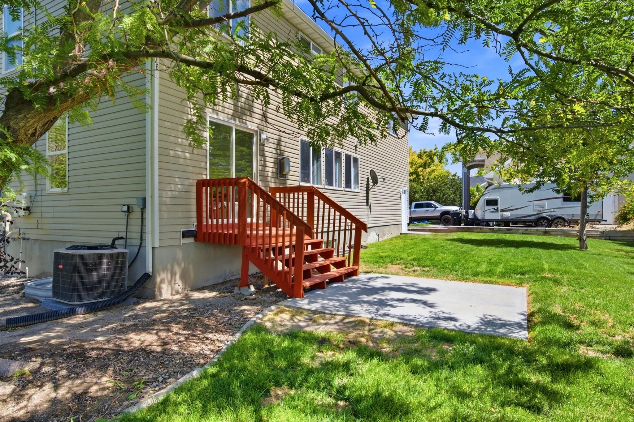 Rear exterior featuring a red-stained wood deck with stairs, a concrete patio, a manicured lawn, and mature trees