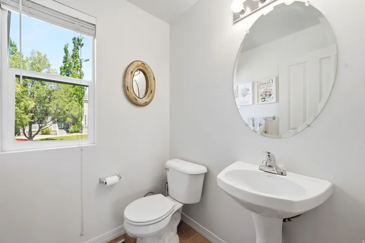 Pedestal sink featuring a chrome faucet, oval mirror, window with privacy blinds, and light tile flooring