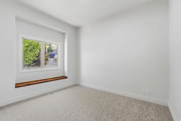 Bright room featuring a window with white trim, a built-in wood-finish shelf, light-toned carpet flooring, crisp white walls, and elegant baseboards