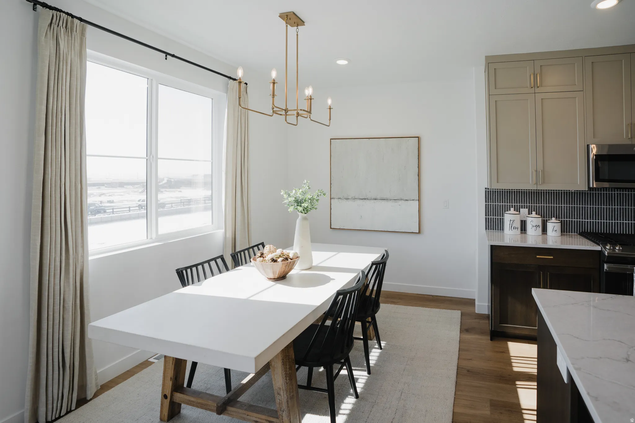 Open-concept dining area featuring a large window with natural light, a rectangular white dining table with a wood base, and a gold linear chandelier