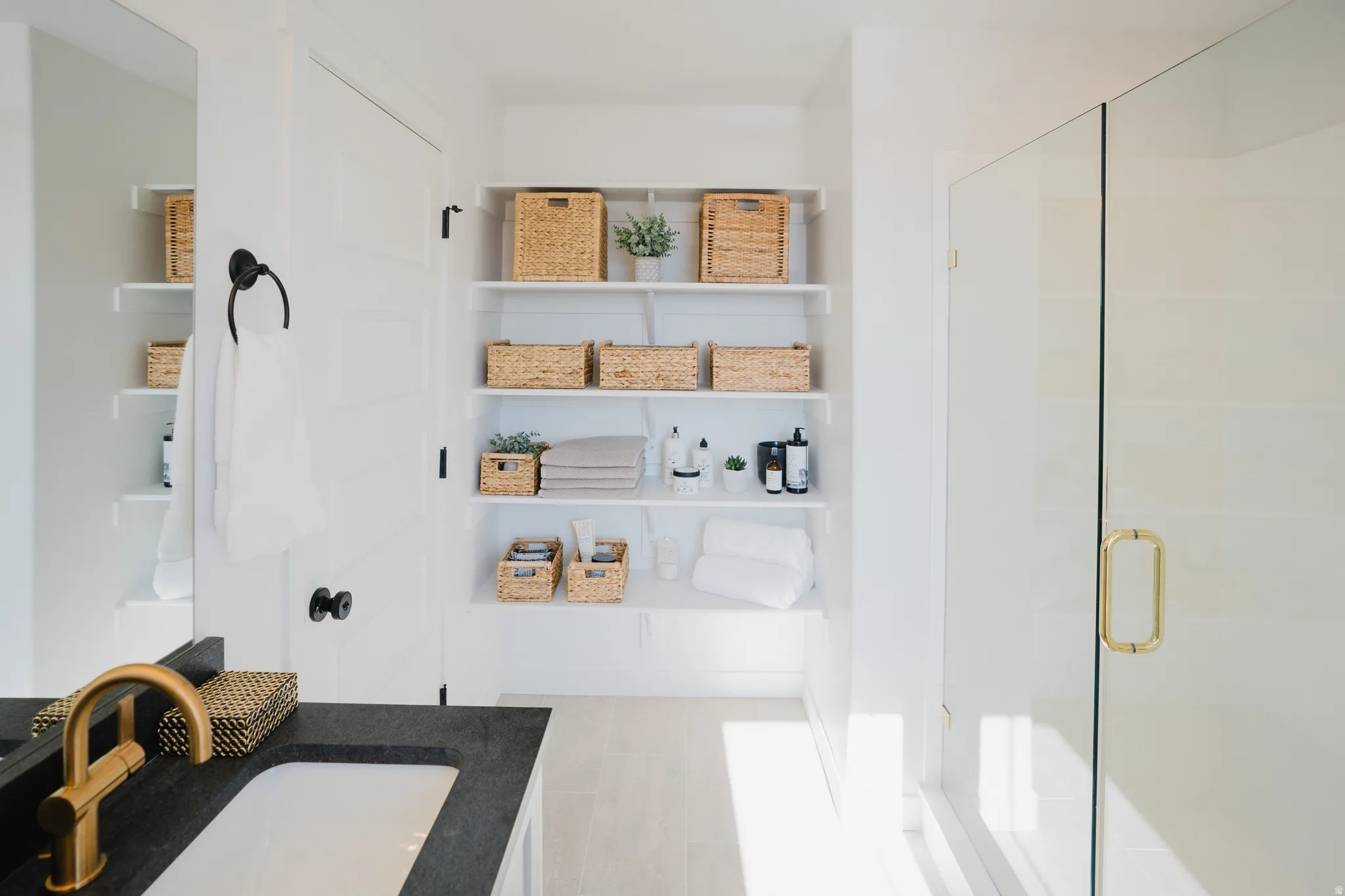 Bathroom featuring a frameless glass shower enclosure with a gold-tone handle, white built-in shelving, a dark countertop vanity with an undermount sink, and a gold-tone faucet