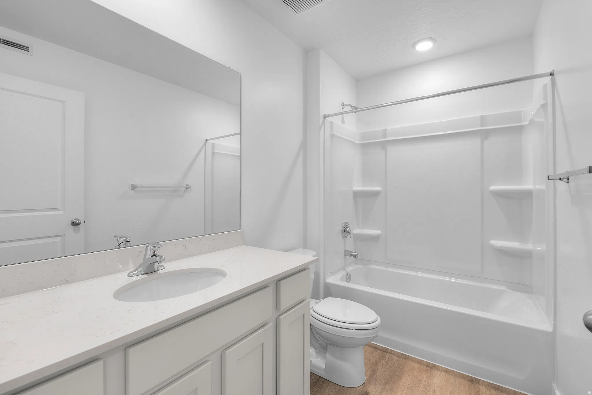 Bathroom featuring a single-sink vanity with a light-toned countertop, a full-width wall mirror, wood-finish flooring, a white toilet, and a white tub/shower combination with integrated shelving